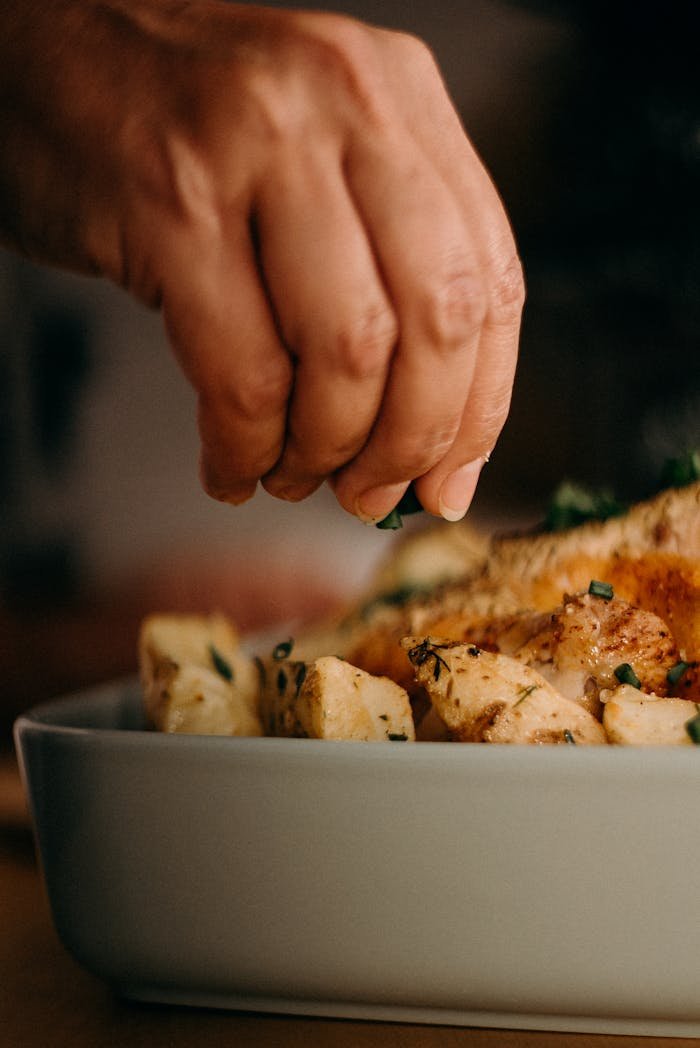 Close-up of a chef's hand garnishing roasted potatoes with herbs, enhancing culinary presentation.